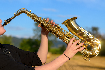 Side close up view of a young woman playing a tenor saxophone outdoors in the countryside raising the instrument in the air against a blue skyの写真素材