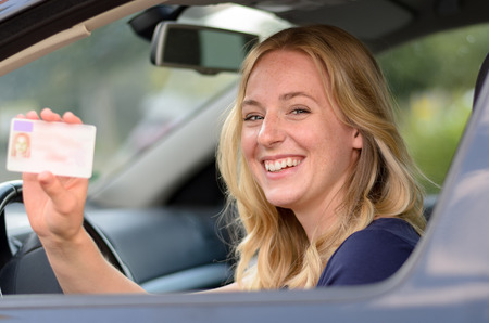 Happy young blond woman sitting behind the steering wheel of a car showing off her drivers license through the open windowの写真素材