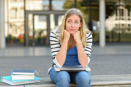 Doubtful female blond teenager with furrowed eyebrows leaning on knees with laptop beside books outside in front of buildingの写真素材