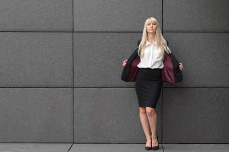 Stylish professional woman opening her suit jacket to reveal her crisp white blouse and shapely body as she leans against a grey tiled commercial wall with copy spaceの写真素材