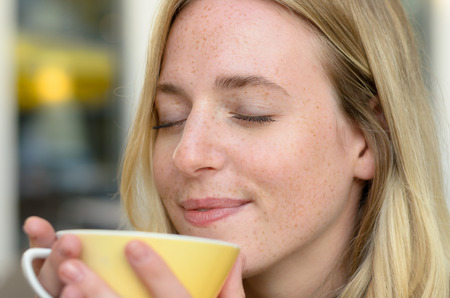 Happy young woman enjoying her morning coffee savoring the aroma with a blissful smile and eyes closed, close up cropped head shotの写真素材