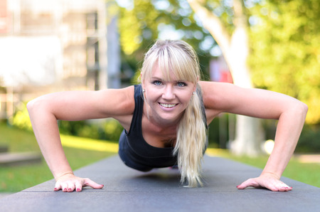 Fit young blond woman doing exercises in a park performing press-ups on a pathway with looking at...