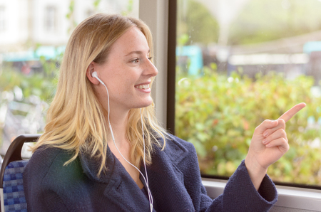 Young attractive blond woman riding in a bus listening to music on earplugs smiling happily as she points to something outside the window in townの写真素材