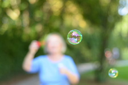 Senior woman blowing iridescent soap bubbles outdoors in a park with, selective focus to the shiny...