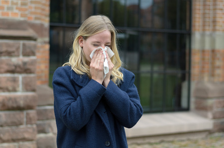 Sick young woman blowing her nose on a tissue or handkerchief as she stands outdoors in town, conceptual of a seasonal flu epidemic or allergic rhinitisの写真素材