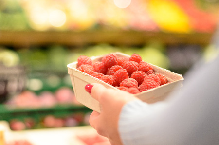 Person selecting a punnet of fresh raspberries to purchase in a supermarket, close up over the shoulder view of the fruitの写真素材