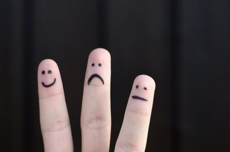 Three different emoticons hand drawn on fingers with a black marker pen with a happy smiling face, unhappy sad face and calm unemotional face isolated on black with copy spaceの写真素材
