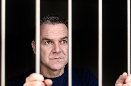 Middle aged blue eyed man incarcerated and wearing blue shirt stands holding prison bars