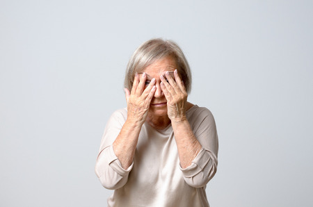 Senior grey-haired woman in light blouse covering her eyes with fingers, both hands to her face, standing against plain grey backgroundの写真素材