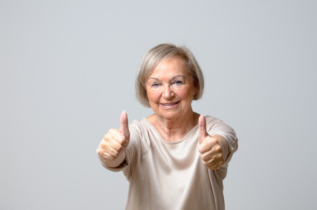 Senior woman in her 70s showing thumbs up gestures with both hands, looking at camera and smiling, standing against plain grey backgroundの写真素材