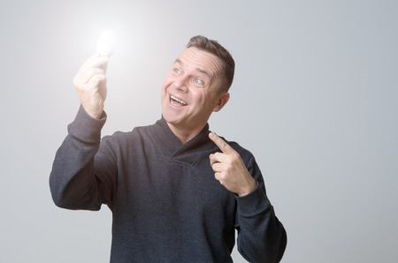 Motivated middle-aged man holding a brightly glowing light bulb with a look of gleeful wonderment conceptual of having a bright idea, breakthrough, inspiration or finding the solution to a problemの写真素材
