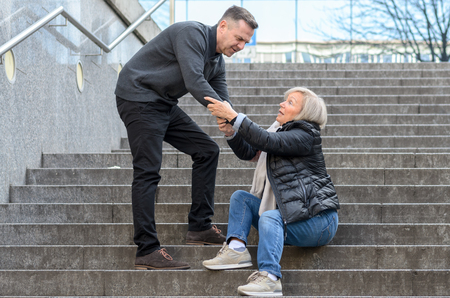 Man helping senior woman to get up while she is sitting on stairs outdoors in the city, holding her kneeの写真素材