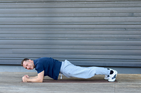 Side view of adult man in training suit doing plank on mat, turning his head to camera and smiling,...
