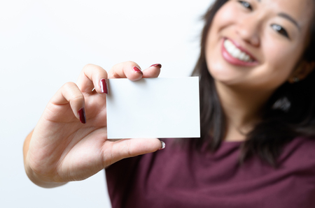 Smiling young Chinese woman holding up a blank white business card on display in a concept of identification, qualifications, contact or branding, focus on cardの写真素材