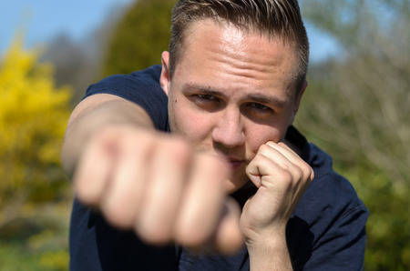Determined young man punching at the camera with his clenched fist with focus to his face and resolute expression outdoors in a gardenの写真素材