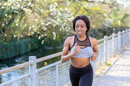 Active athletic young African woman with a muscular physique out jogging along a country road past...