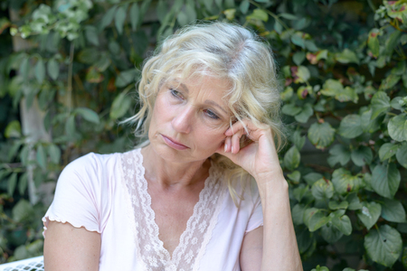 Despondent middle-aged woman sitting thinking outdoors in front of greenery with her head on her hand staring off morosely to the sideの写真素材