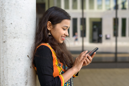 Happy attractive friendly young Indian student with long hair texting on her mobile phone as she leans against a wall outdoorsの写真素材