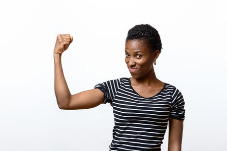 Cute young African woman flexing her muscles with a mischievous look as she raises her fist and arm isolated over whiteの写真素材