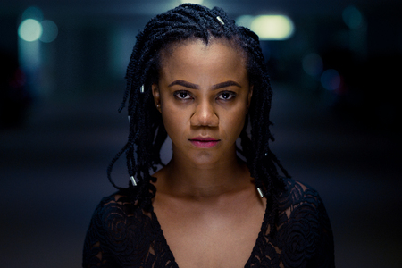 Night portrait of a serious stylish attractive young African woman with braided hair with shining end adornments in a shadowy interior space with defocused lightsの写真素材