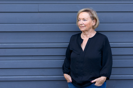 Friendly relaxed senior woman in a chic trendy outfit standing against a blue background with her...