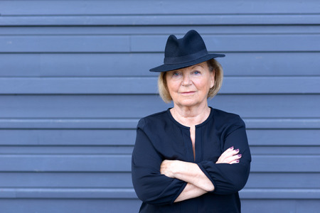 Confident trendy senior lady in a stylish hat standing with folded arms over a blue background with parallel lines gazing at the camera with a thoughtful expressionの写真素材
