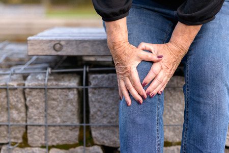 Elderly lady grabbing her knee in pain as she sits on a wall surrounding a pond after injuring herself in a close up view.の写真素材