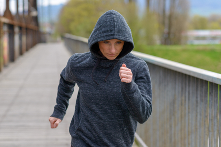 Athletic woman in a hoodie jogging across a bridge approaching the camera with a look of concentration in a health and fitness conceptの写真素材