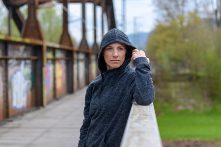 Serious thoughtful woman standing on an old iron bridge leaning on the parapet lifting her hood on her sweater to the side with her handの写真素材