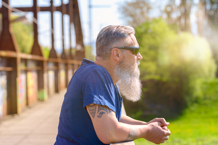 Bearded grey-haired man in sunglasses standing on an old bridge leaning on the railing, side viewの写真素材
