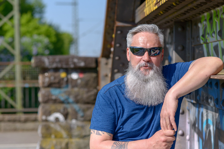 Trendy attractive bearded grey-haired man in sunglasses standing leaning against a girder on an old bridge in the sunshine looking at the cameraの写真素材