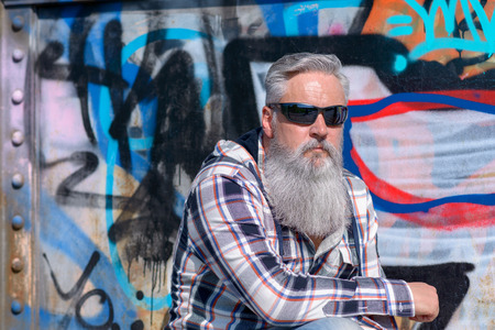Mature man with long grey beard wearing sunglasses against painted wall on sunny dayの写真素材