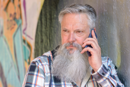 Thoughtful grey-haired bearded man listening to a mobile phone call as he stands in front of a wallの写真素材