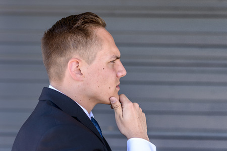 Thoughtful young businessman in profile standing with his hand to his chin as he stares ahead against a grey wallの写真素材