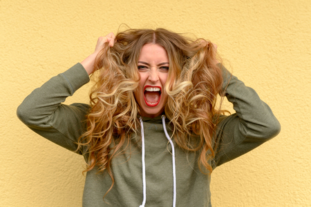 Frustrated blond woman tearing at her long wavy hair and yelling to vent her feelings over a yellow studio backgroundの写真素材