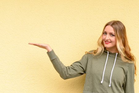 Smiling attractive blond woman holding out her empty hand for product placement over blank copy space over a yellow studio backgroundの写真素材