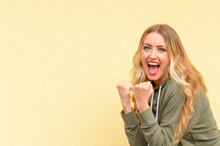 Excited blond woman cheering and clenching her fists as she celebrates a personal victory or success over a yellow studio background with copy spaceの写真素材