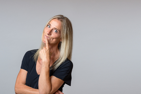 Speculative woman looking at the camera with wide eyed thoughtful expression with her head tilted to the side and hand to her chin over grey with copy spaceの写真素材