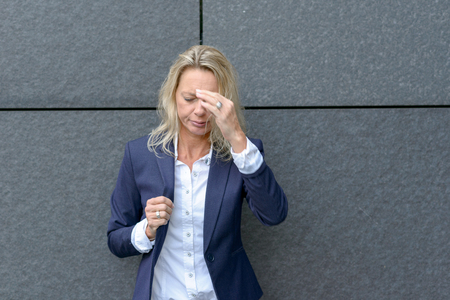 Stylish professional woman suffering from a headache standing holding her hand to her forehead in front of a grey wall looking down with a pained expressionの写真素材