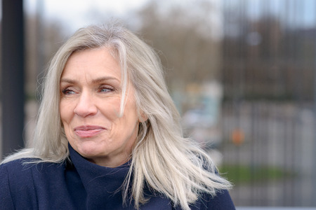 Attractive mature woman with tousled shoulder length blond hair looking to the left of the frame with a pleased smile against a blurred urban backgroundの写真素材