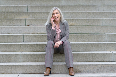 Happy businesswoman using her mobile in town sitting on a flight of steps smiling as she chats on the callの写真素材