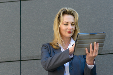 Mature businesswoman using a handheld tablet to surf the internet as she pauses in front of a grey stone wall in townの写真素材