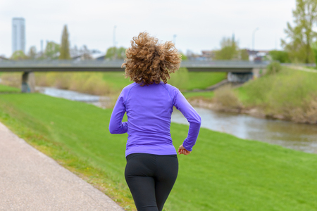 Athletic woman in sportswear out jogging alongside a rural canal or river running away from the camera with her hair blowing behind her in a fitness and healthy lifestyle conceptの写真素材