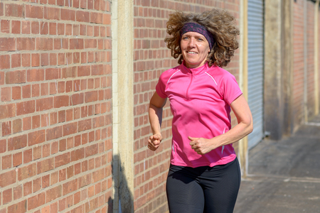 Healthy middle-aged woman jogging through town on the sidewalk past brick walls in colorful pink sportswear in a fitness and active lifestyle conceptの写真素材