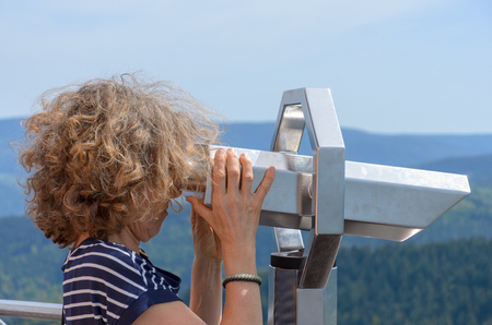Woman looking through a binocular telescope while on vacation in the Black Forest region in Germany in a close up side view on a mountain viewpointの写真素材