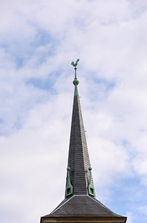 Old historic tall tiled spire on a roof surmounted by the figure of a cock against a cloudy blue skyの写真素材