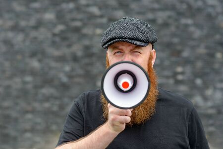 Bearded man wearing a cloth cap speaking into a megaphone making an announcement or public speaking