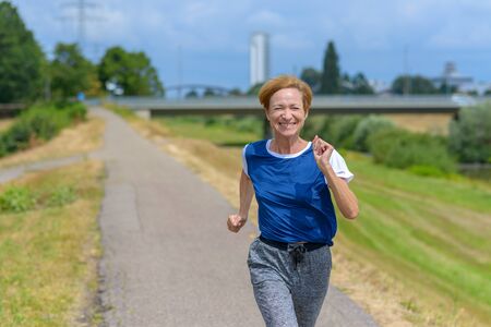 Fit athletic middle-aged woman out jogging along a rural road smiling as she approaches the cameraの写真素材