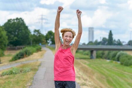 Exuberant vivacious woman celebrating outdoors laughing and raising her arms in the air on a quiet country roadの写真素材