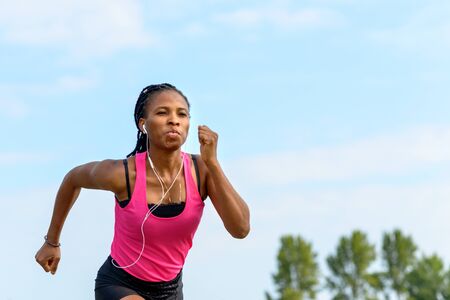 Fit determined young African woman jogging while listening to her music on ear buds passing the camera in a low angle view in a health and fitness conceptの写真素材